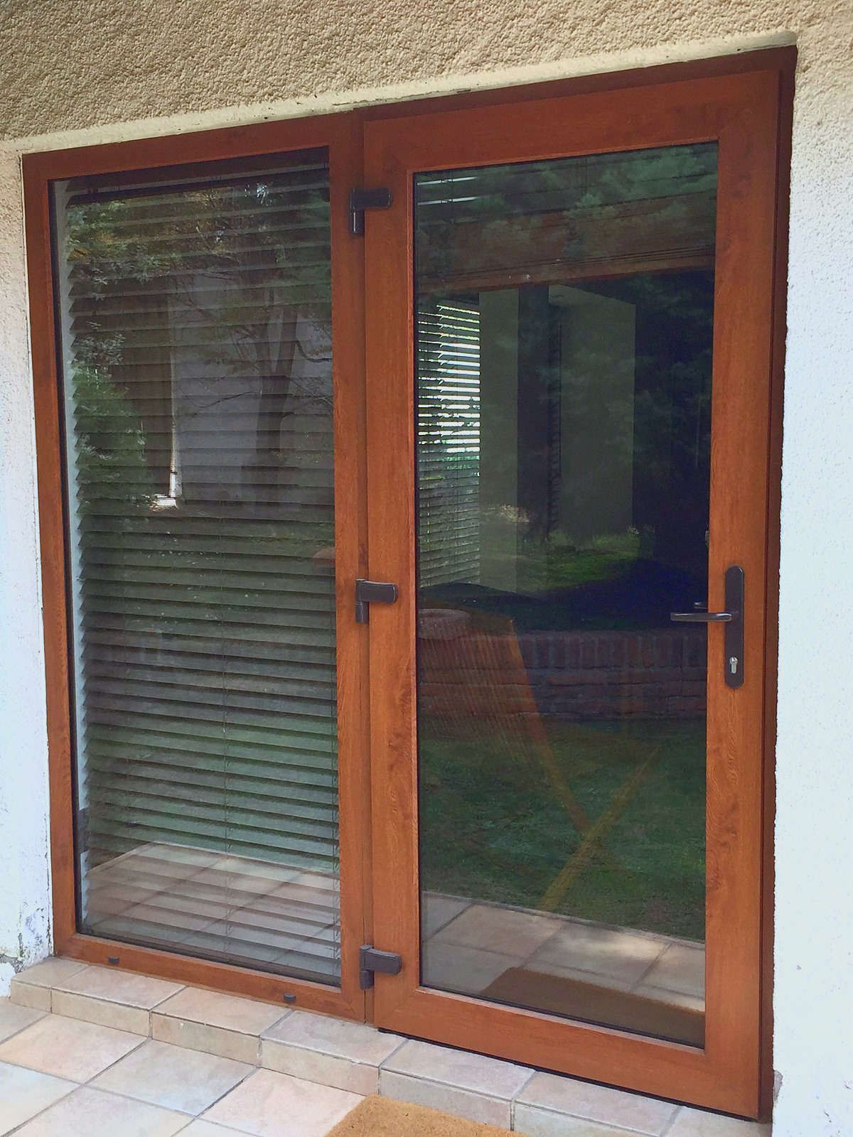Puerta abatible de dos hojas, de PVC foliado imitación nogal, con bisagras y manilla negras, hacia un patio con pasto. Es PVC foliado imitación madera, no es madera.