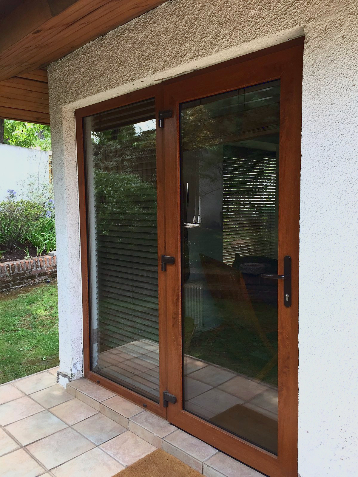 Puerta ventana de PVC foliado imitación nogal, con manilla negra, en un muro blanco junto al jardín. Es PVC foliado imitación madera, no es madera.