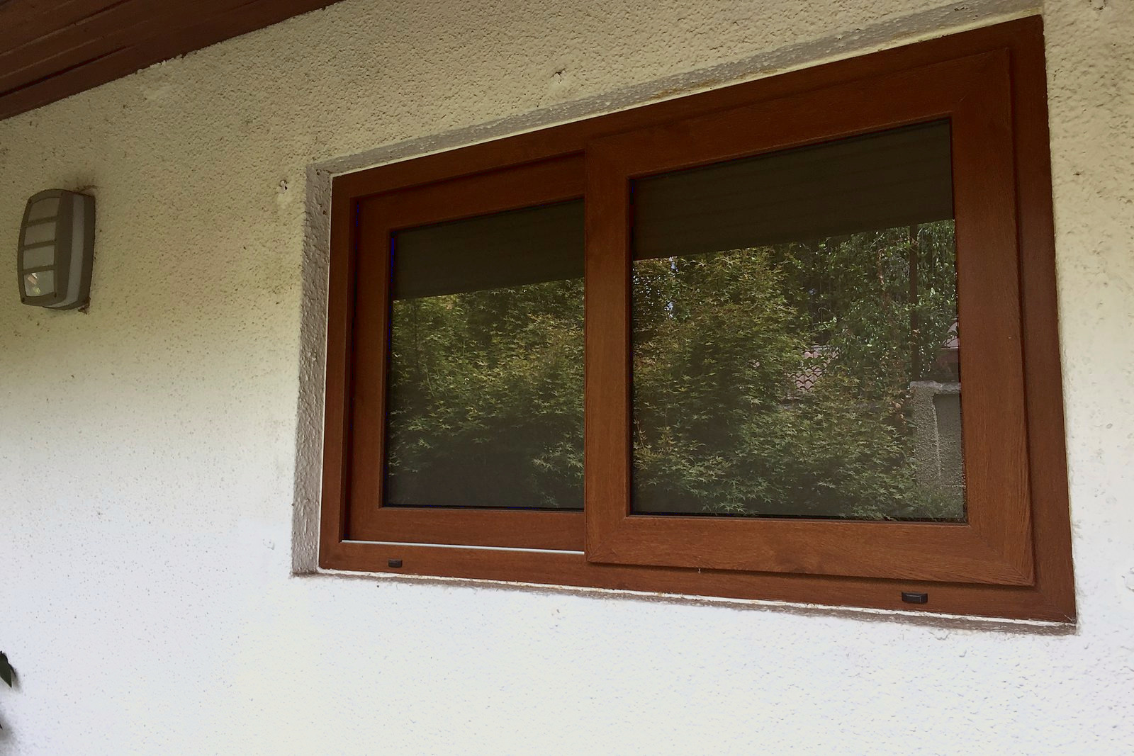 Ventana corredera de dos hojas, de PVC foliado imitación nogal, en un muro exterior blanco, con persiana interior. Es PVC foliado imitación madera, no es madera.