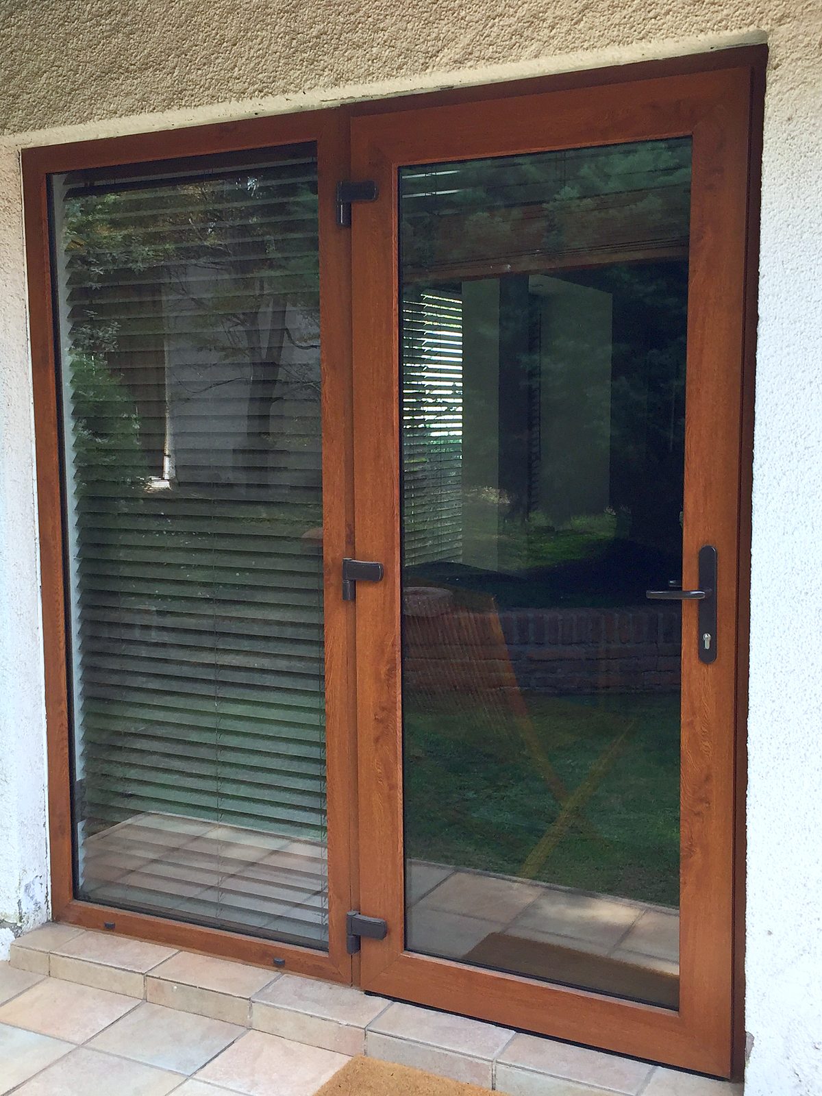 Puerta abatible de PVC foliado imitación madera nogal, de dos hojas, con bisagras y manilla negras, que da a un patio con pasto. No es madera.