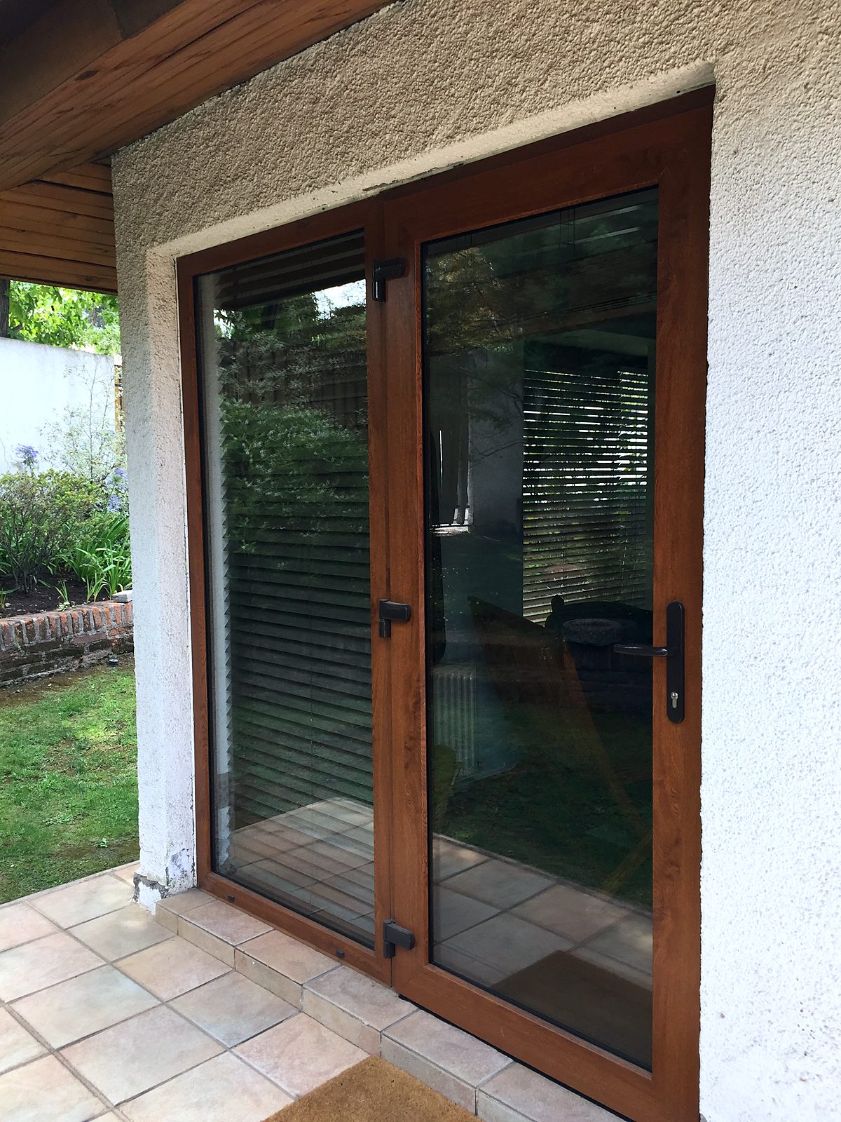 Puerta ventana de PVC foliado imitación madera nogal, con manilla negra, instalada en un muro blanco junto al jardín. No es madera.