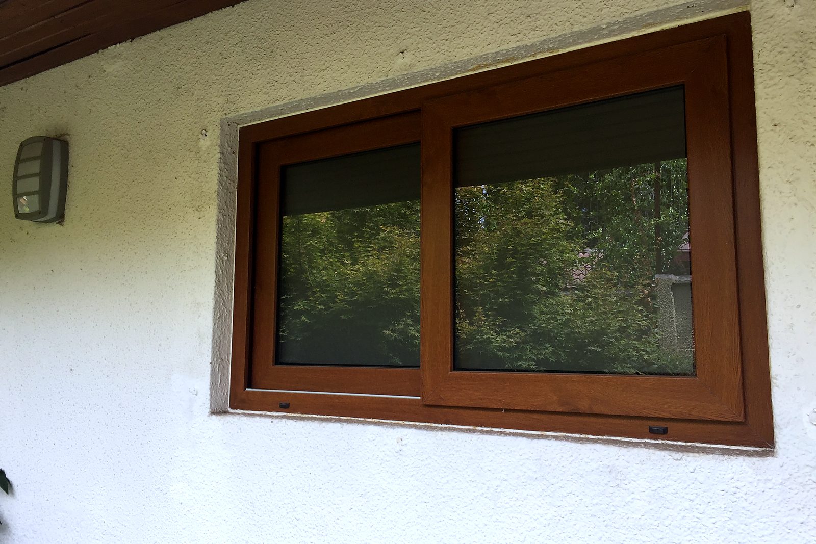 Ventana corredera de PVC foliado imitación madera nogal, de dos hojas, instalada en un muro exterior blanco, con persiana interior. No es madera.